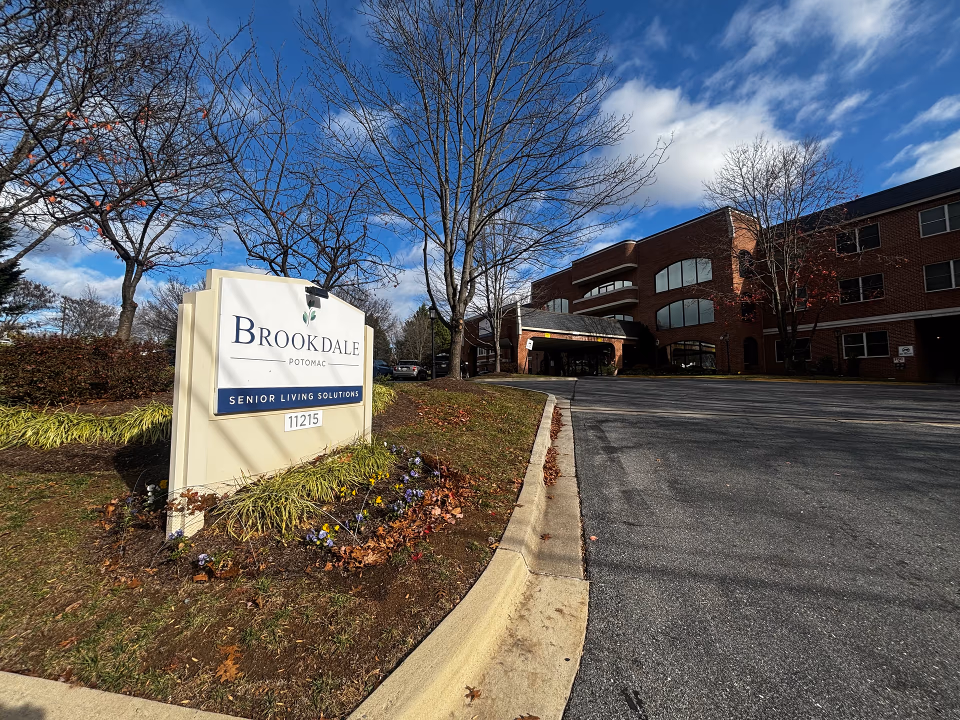 Exterior view of the Brookdale Potomac senior living facility with a large sign in the foreground displaying the facility name and address. The building is a multi-story brick structure with large windows and a covered entrance. Trees with sparse leaves and a partly cloudy blue sky are visible.