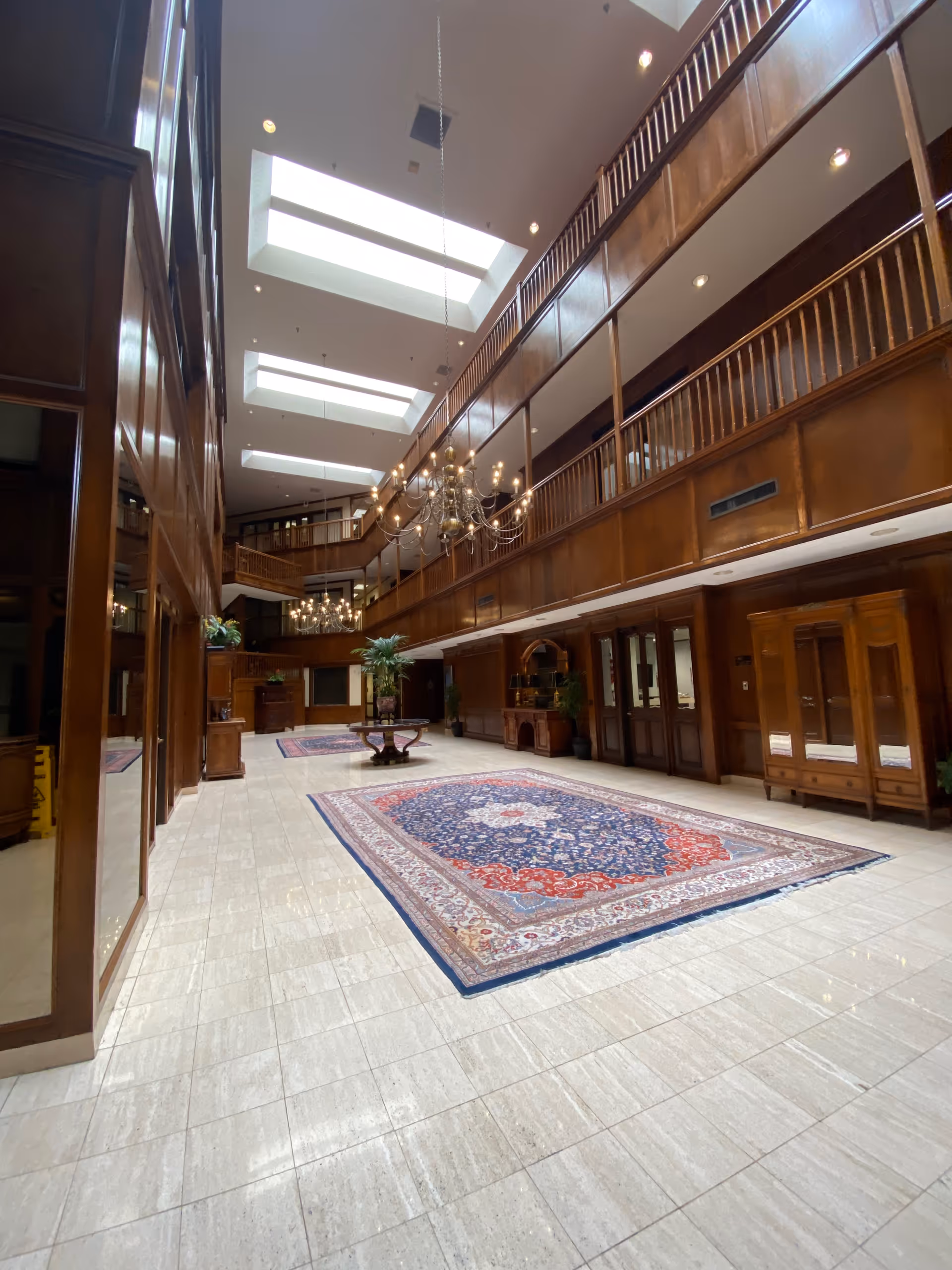 A spacious, wood-paneled senior living lobby with skylights, chandeliers, and a large central rug.