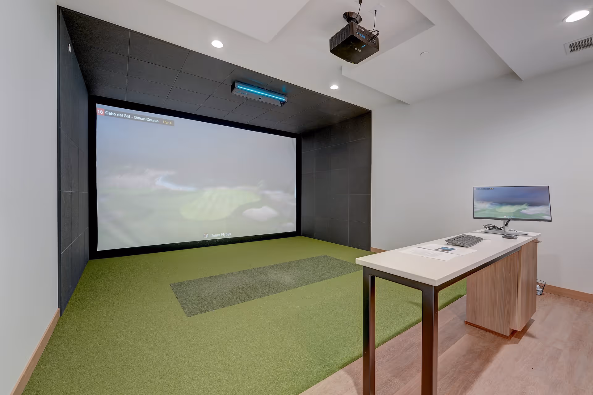Indoor golf simulator room with a large projection screen, green turf floor, and a desk with a computer monitor.