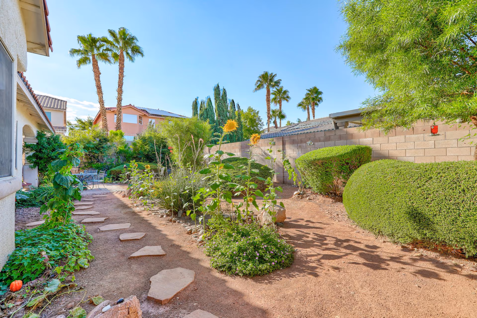 Sunny outdoor garden with stepping-stone path, shrubs, sunflowers, palm trees, and a brick wall.