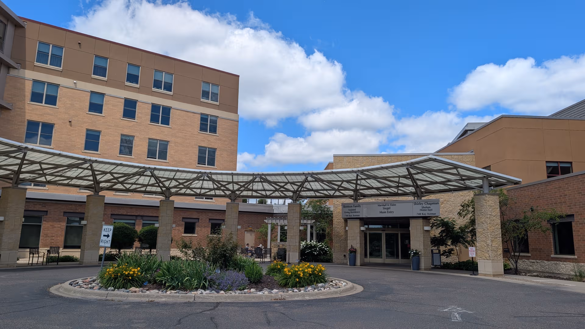 Front view of a senior living facility entrance with a glass canopy and landscaped roundabout.
