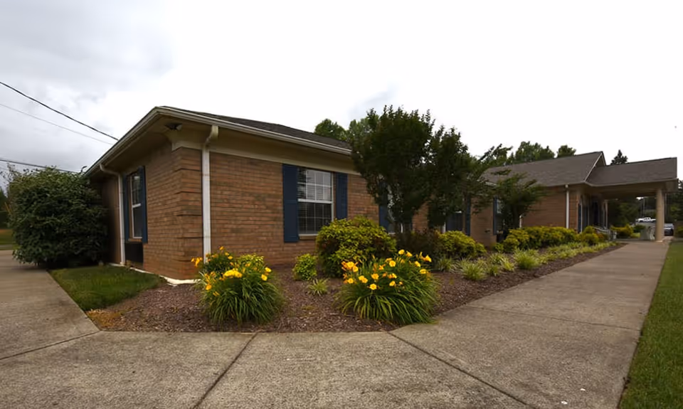 Exterior view of a single-story brick building with blue window shutters, surrounded by landscaped flower beds with yellow flowers and green shrubs, and a concrete walkway leading to the entrance under a covered porch.