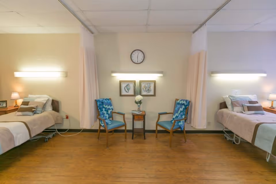 Shared patient room with two beds separated by privacy curtains, two chairs and a small table under a wall clock.