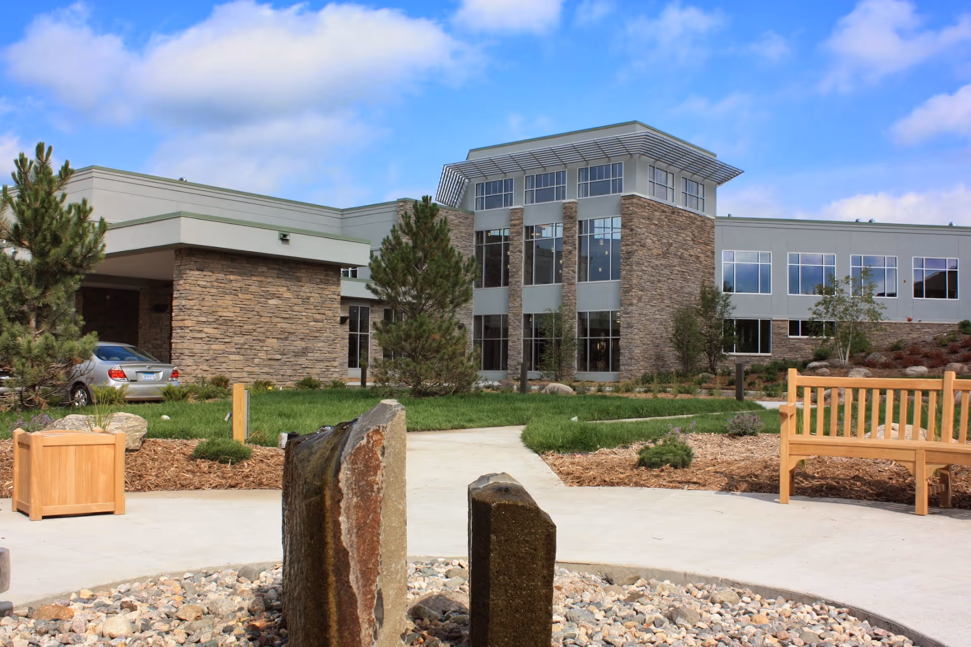Exterior view of a modern senior living facility building with stone and glass facade, surrounded by landscaped greenery, trees, a wooden bench, and a small rock water feature in the foreground under a blue sky with some clouds.