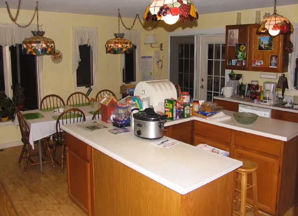 A kitchen and dining area with wooden cabinets and a white countertop island. The kitchen has various appliances including a slow cooker, toaster oven, coffee maker, and blender. There are three stained glass pendant lights hanging from the ceiling. The dining area has a table covered with a white tablecloth and six wooden chairs. The walls are painted yellow with three windows covered by white curtains. There are various food items and kitchen supplies on the countertops.
