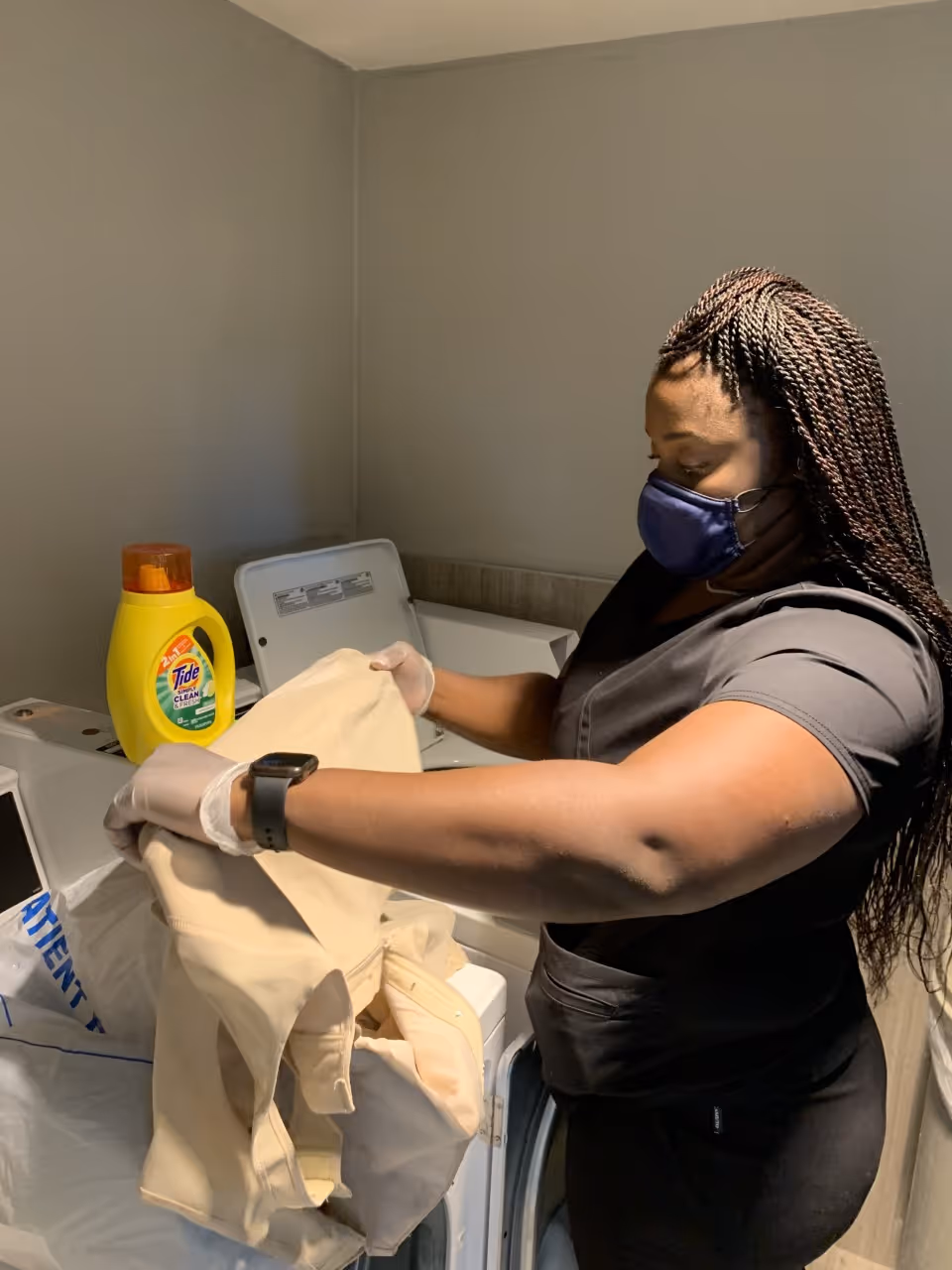 Caregiver wearing a mask and gloves loads clothes into a washing machine in a laundry room.