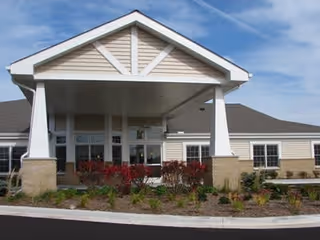 Front exterior view of a single-story assisted living facility building with a covered entrance supported by white pillars, beige siding, and landscaped bushes and plants in front.
