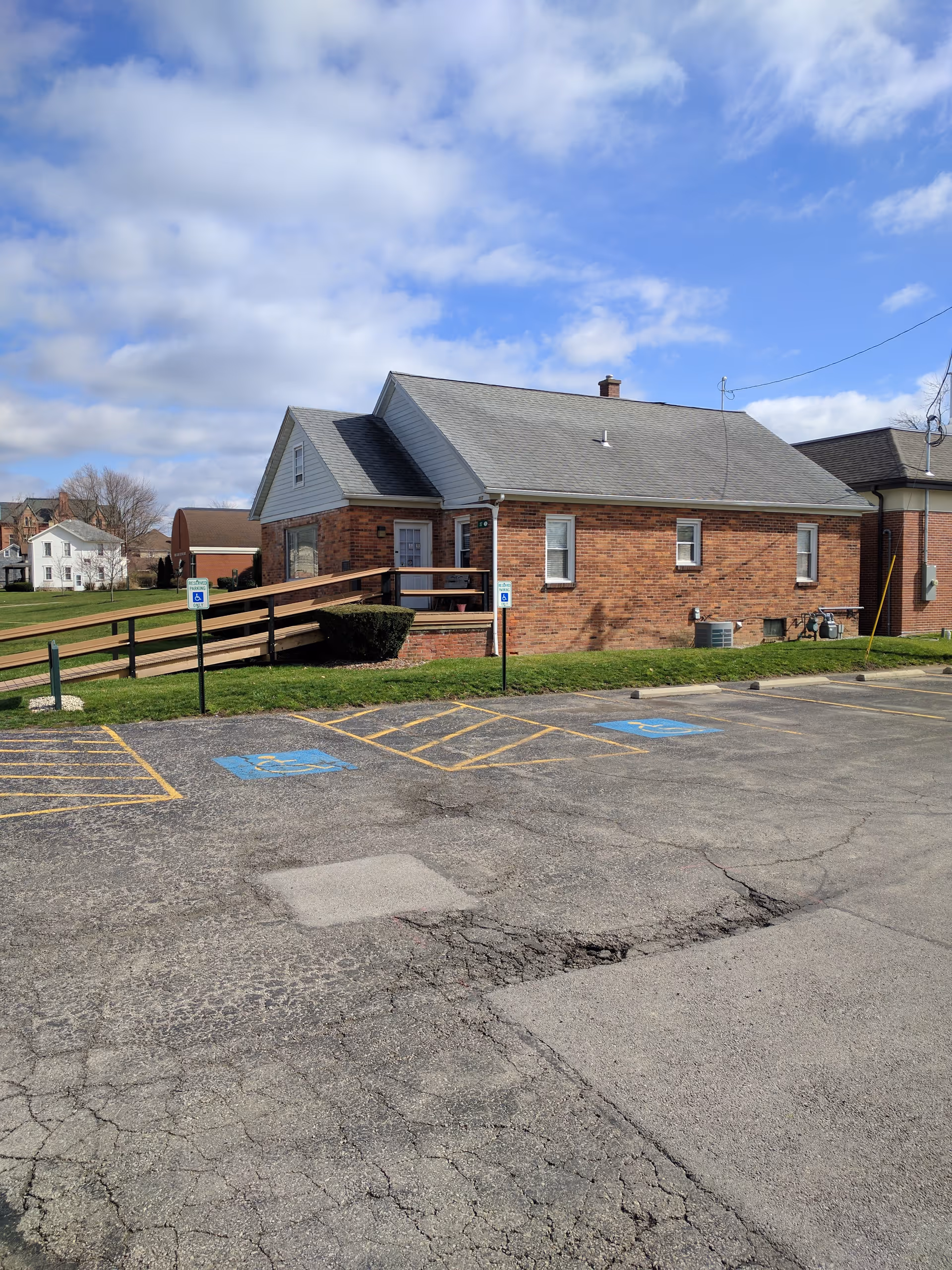 Brick senior living facility with front entrance, ramp, and handicap parking spots in a sunny lot.