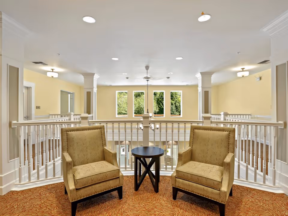Two beige upholstered armchairs and a small round table in a bright upstairs common seating area overlooking an open atrium with windows.
