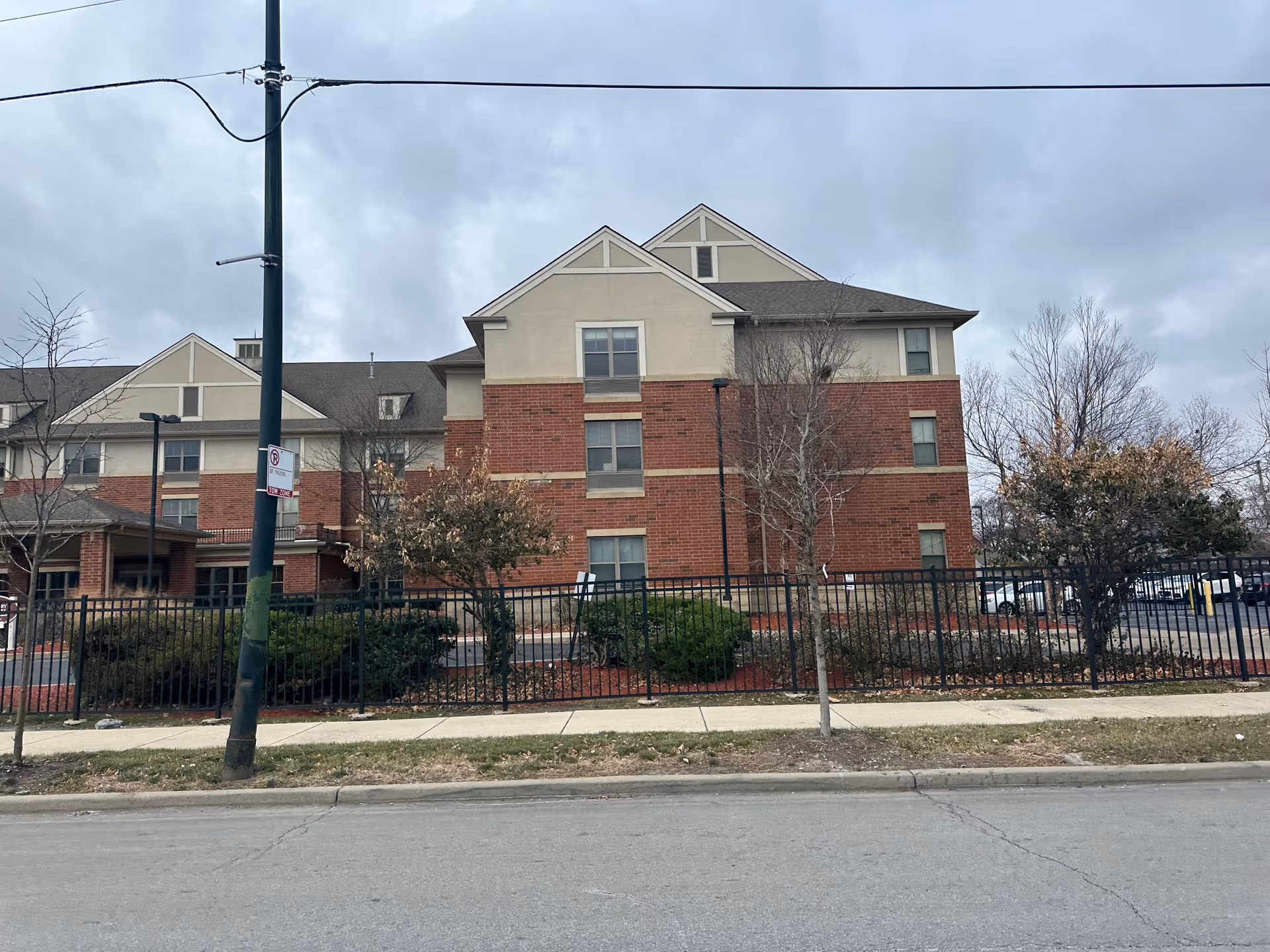 Front exterior of a three-story brick and stucco senior living building behind a black metal fence and sidewalk.