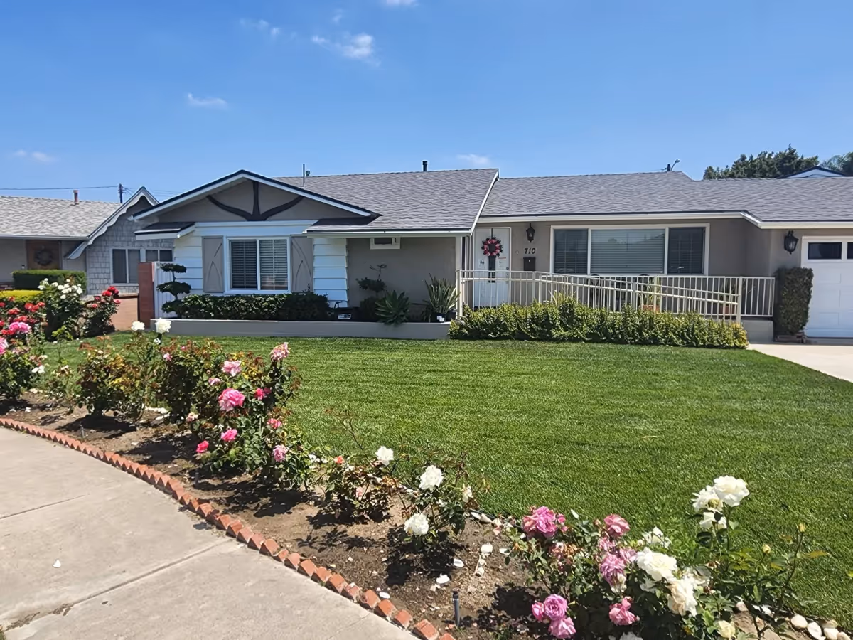 Front exterior view of a single-story residential building with a well-maintained green lawn and a flower bed with pink and white roses along the sidewalk. The house has a ramp leading to the front door, which is decorated with a wreath. The sky is clear and blue.