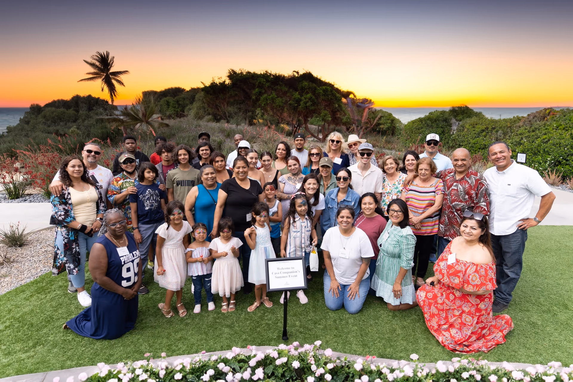 Large diverse group posing outdoors on a lawn during sunset.