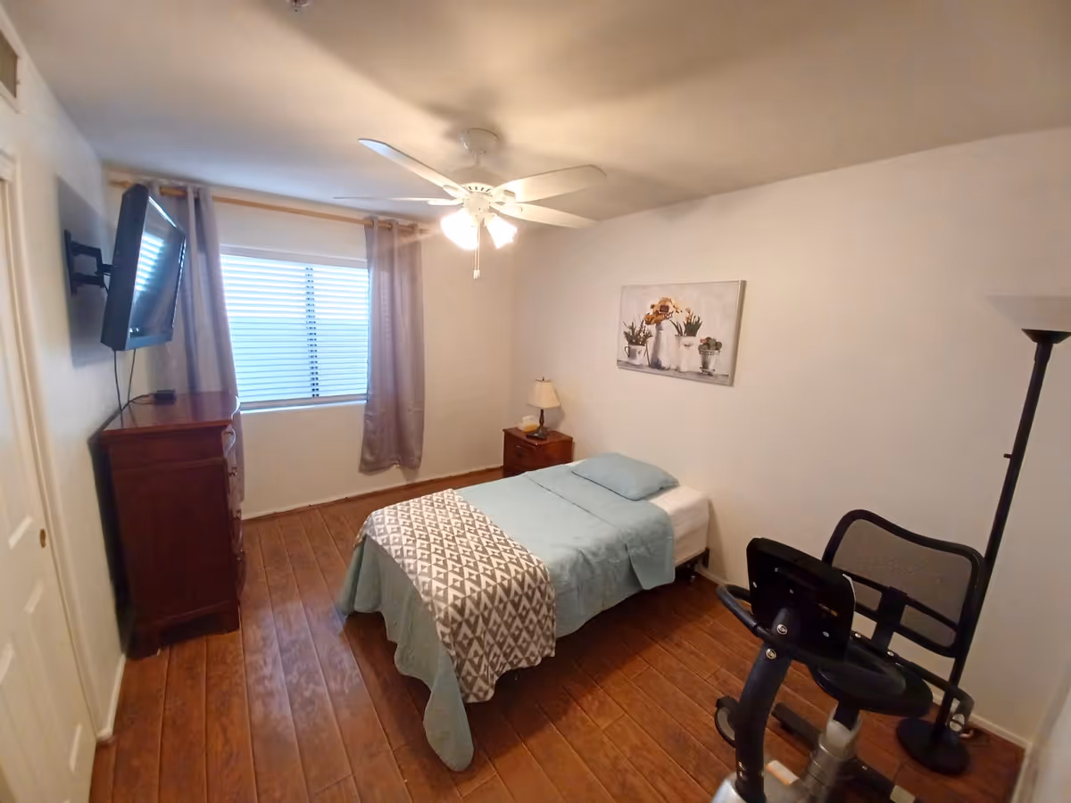 Cozy bedroom with bed, dresser, window, ceiling fan, lamp, and exercise bike.