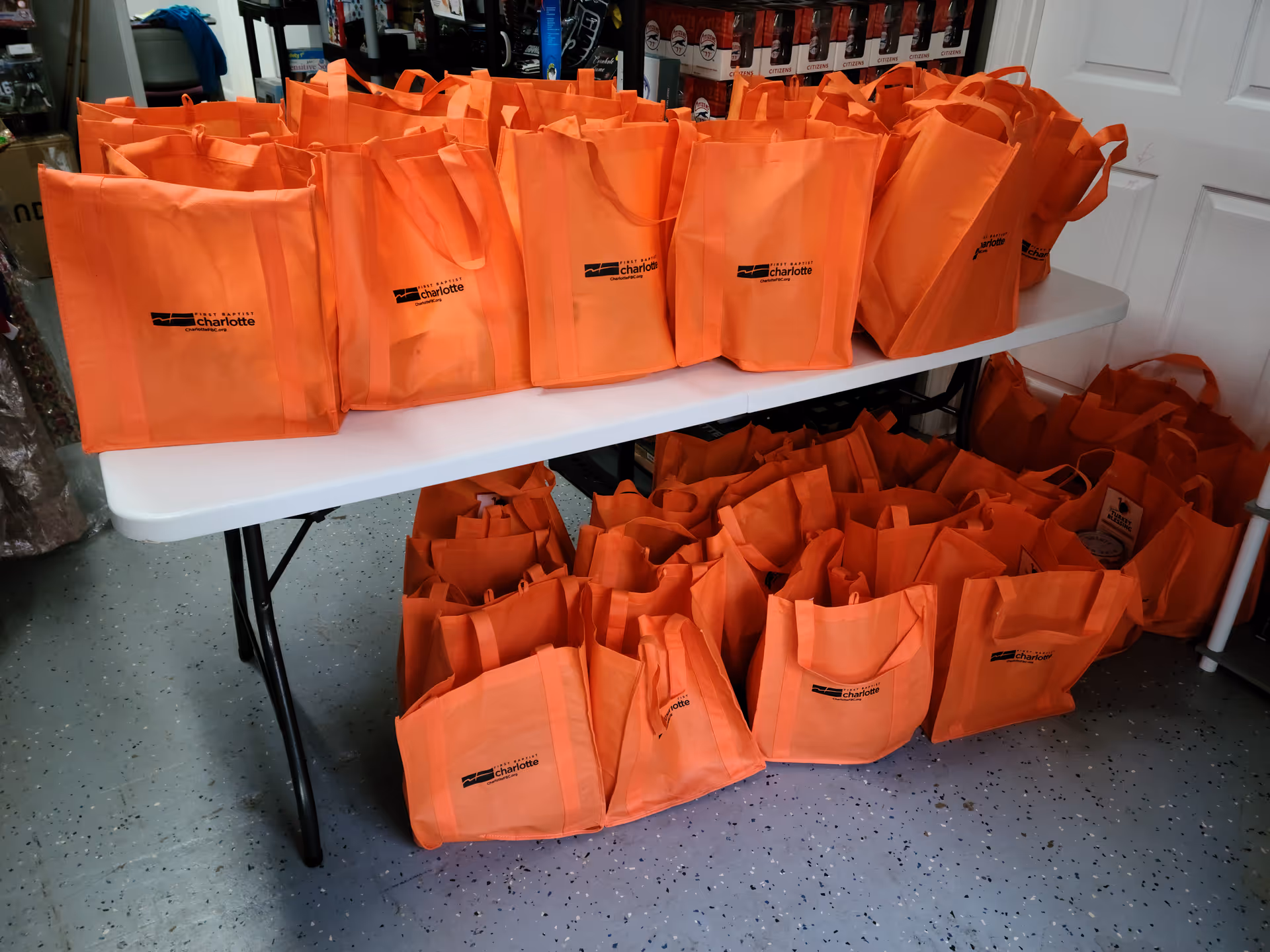 Many orange tote bags with logos arranged on a folding table in a storage room.
