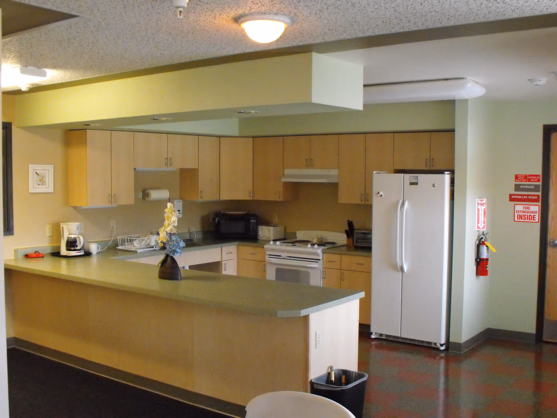 Interior view of a kitchen area with light wood cabinets, a white refrigerator, a white stove, a microwave, a coffee maker, and a countertop with a vase of flowers. There is a fire extinguisher mounted on the wall near a door, and the floor has a checkered pattern.