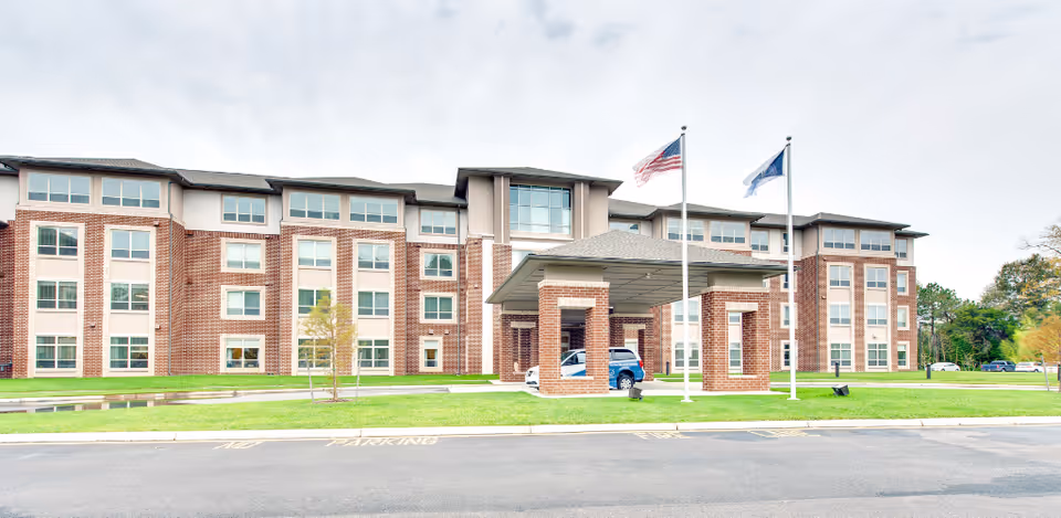 Brick senior living facility front with porte-cochere, flags, and manicured lawn.