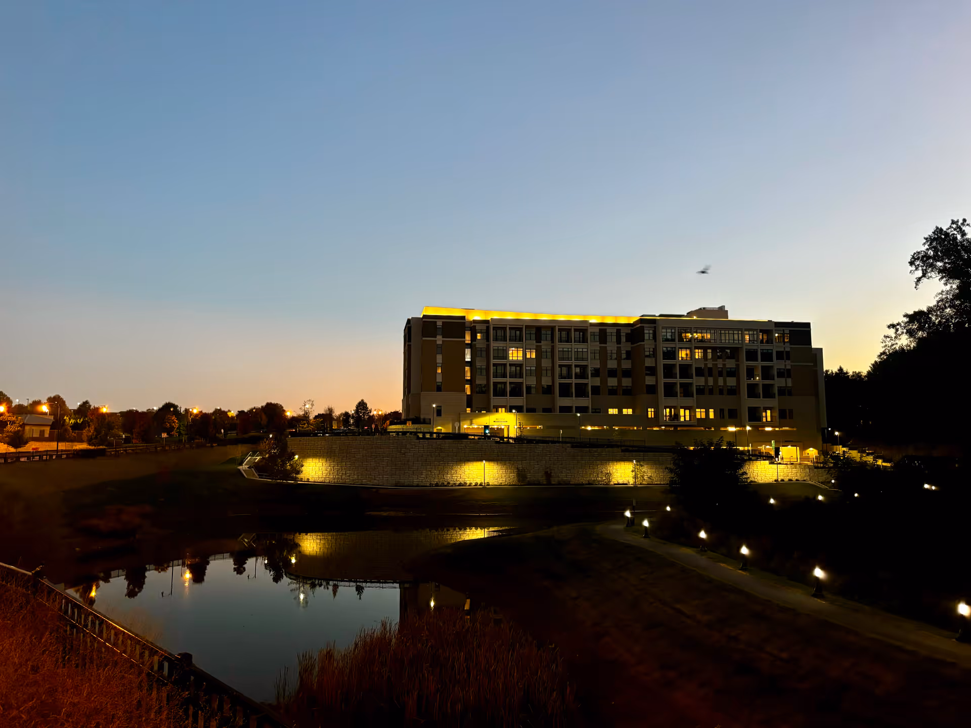 Large multi-story senior living building lit at dusk with a reflecting pond and landscaped path in the foreground.