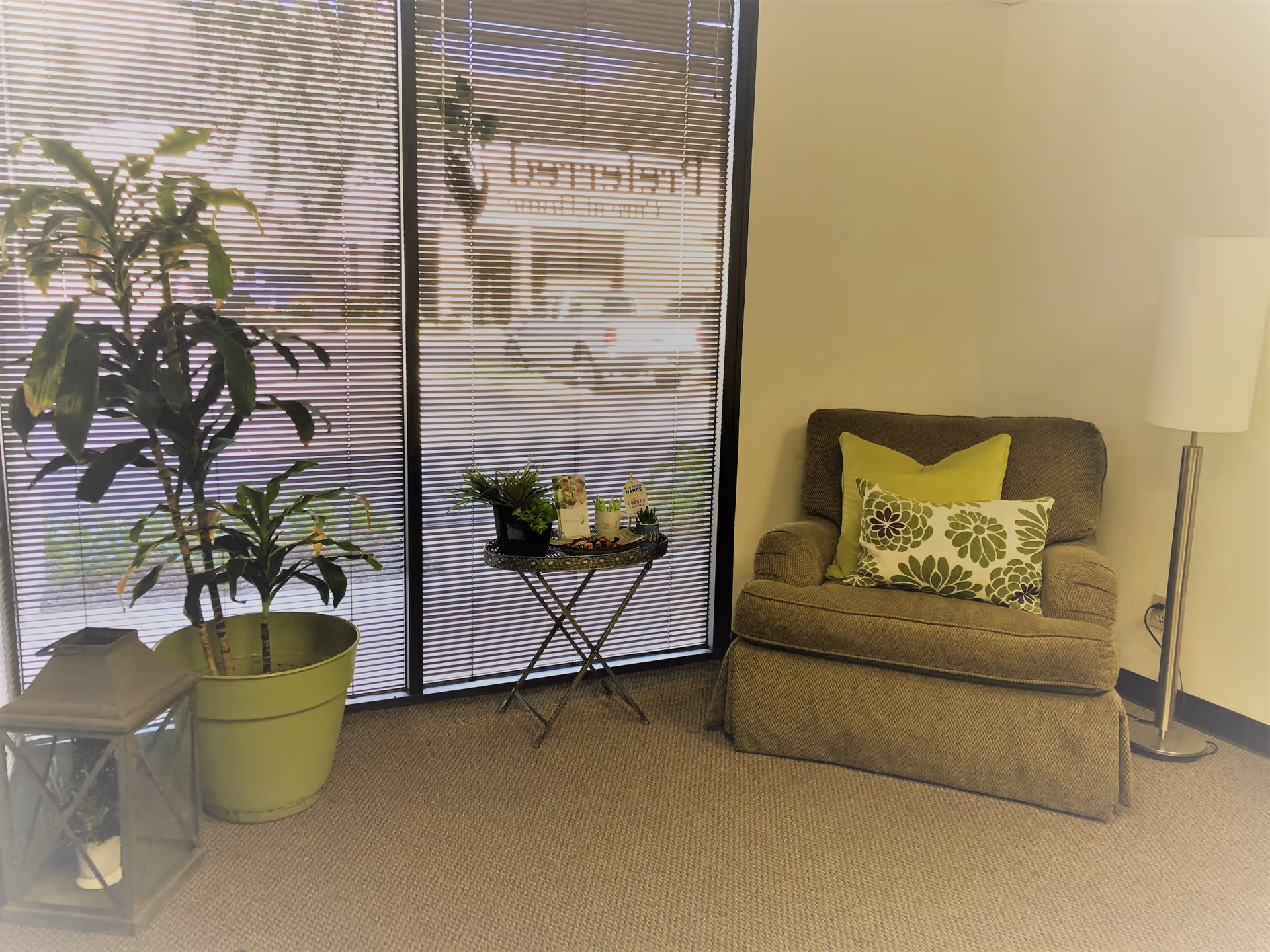 Interior living room area with a beige armchair, plants, blinds, and lamp.