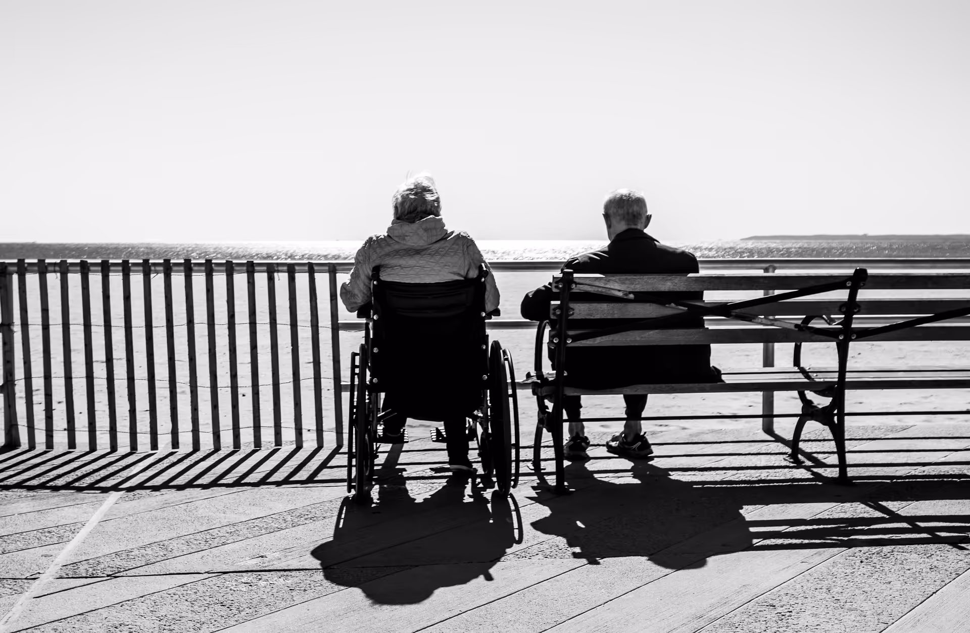 Two elderly people sit on a seaside boardwalk, one in a wheelchair, facing the water.