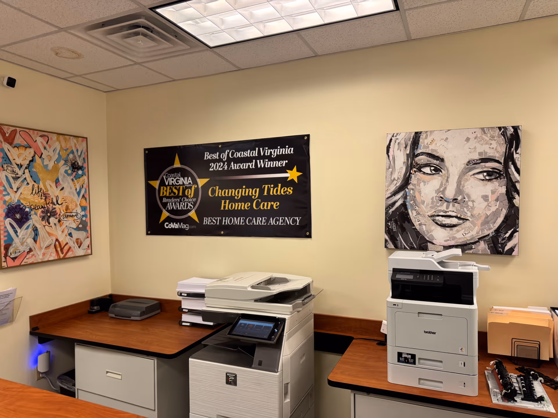 Office workspace with two printers, a banner reading Changing Tides Home Care, and wall art.