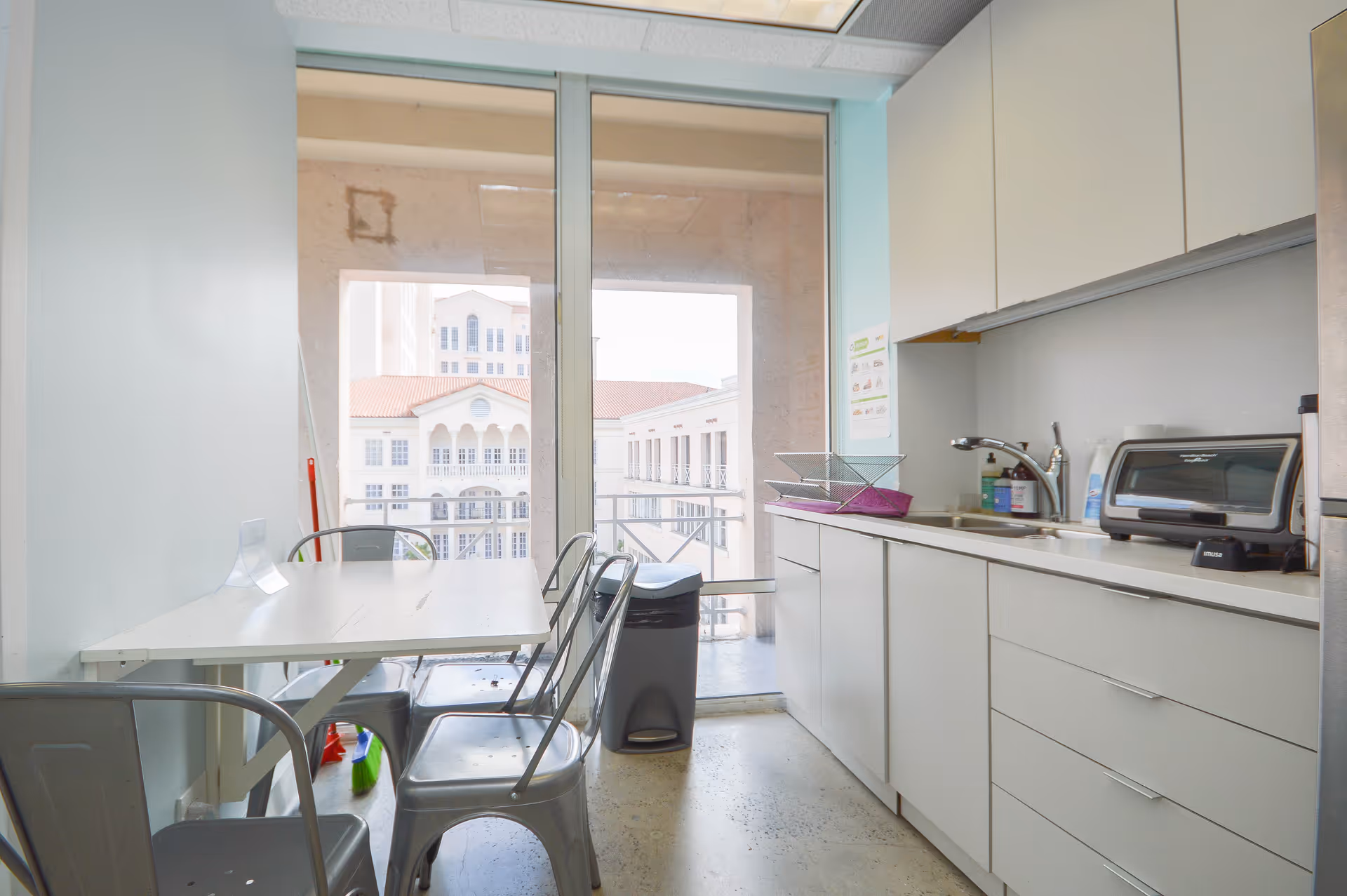 Bright kitchenette with white cabinets, sink, toaster, microwave, and a small table with metal chairs.