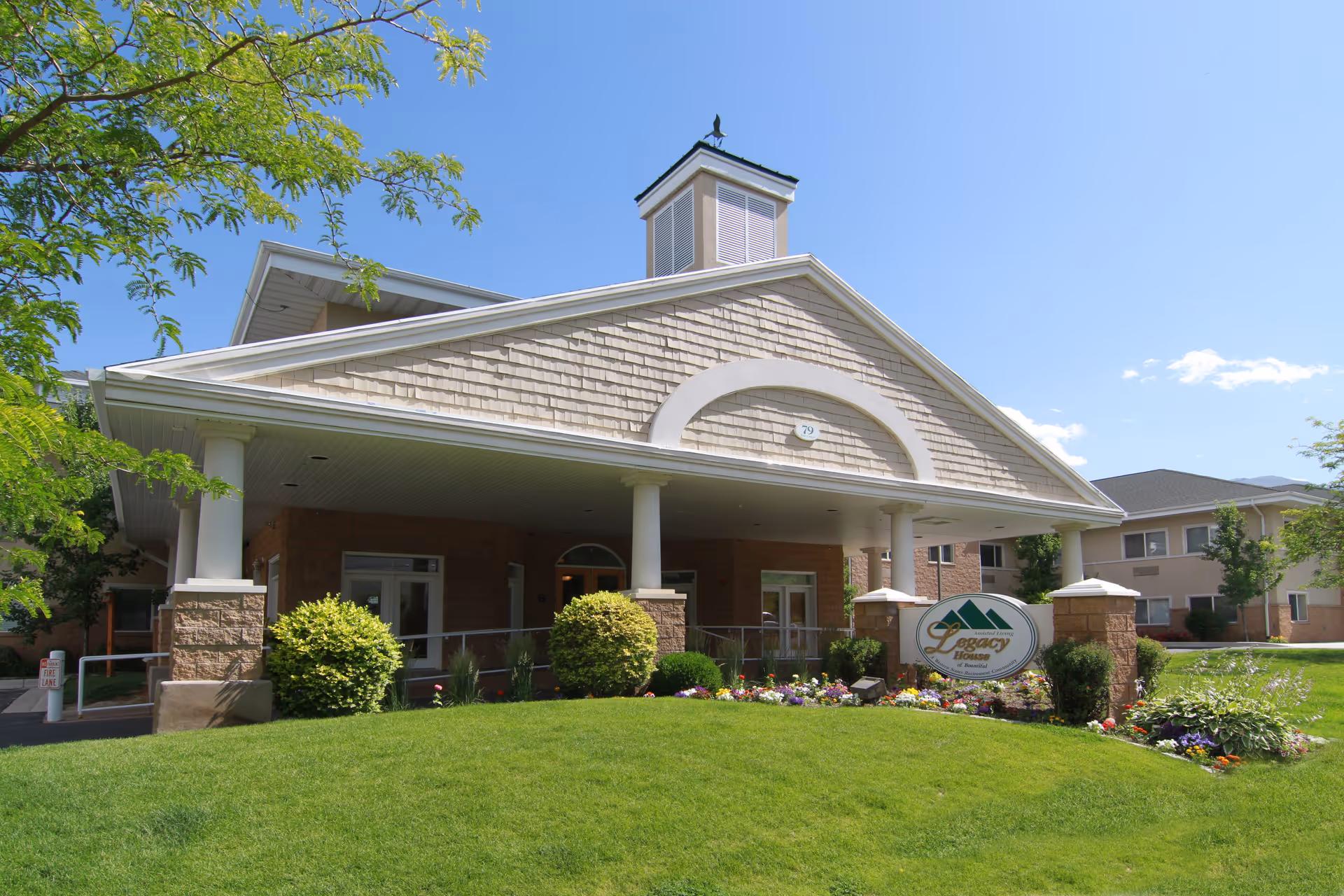 Exterior front view of Legacy House of Bountiful, a senior living facility with a covered entrance supported by white columns, surrounded by green grass, bushes, and colorful flowers under a clear blue sky.