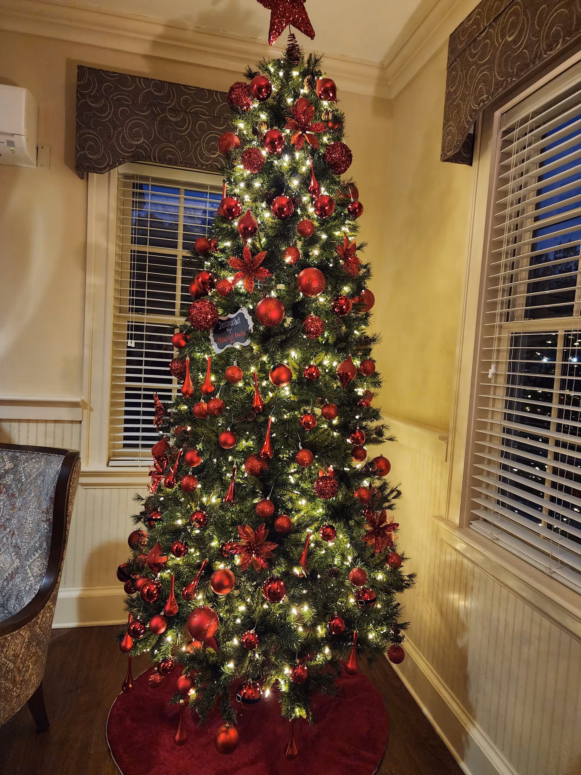 A festively decorated Christmas tree stands indoors beside windows in a cozy room.