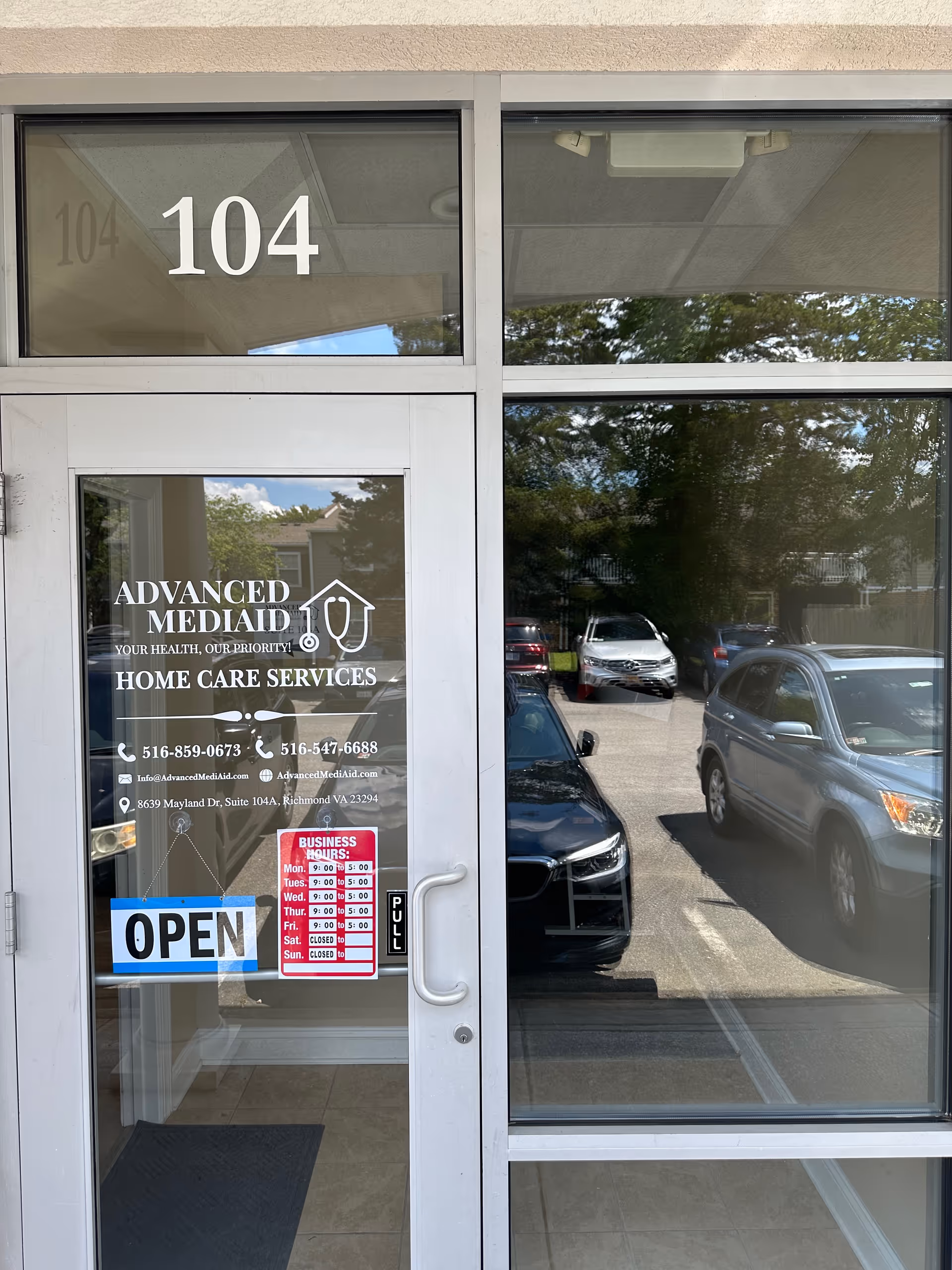 Glass front door with Advanced MediAid signage, OPEN sign, and reflected parked cars.