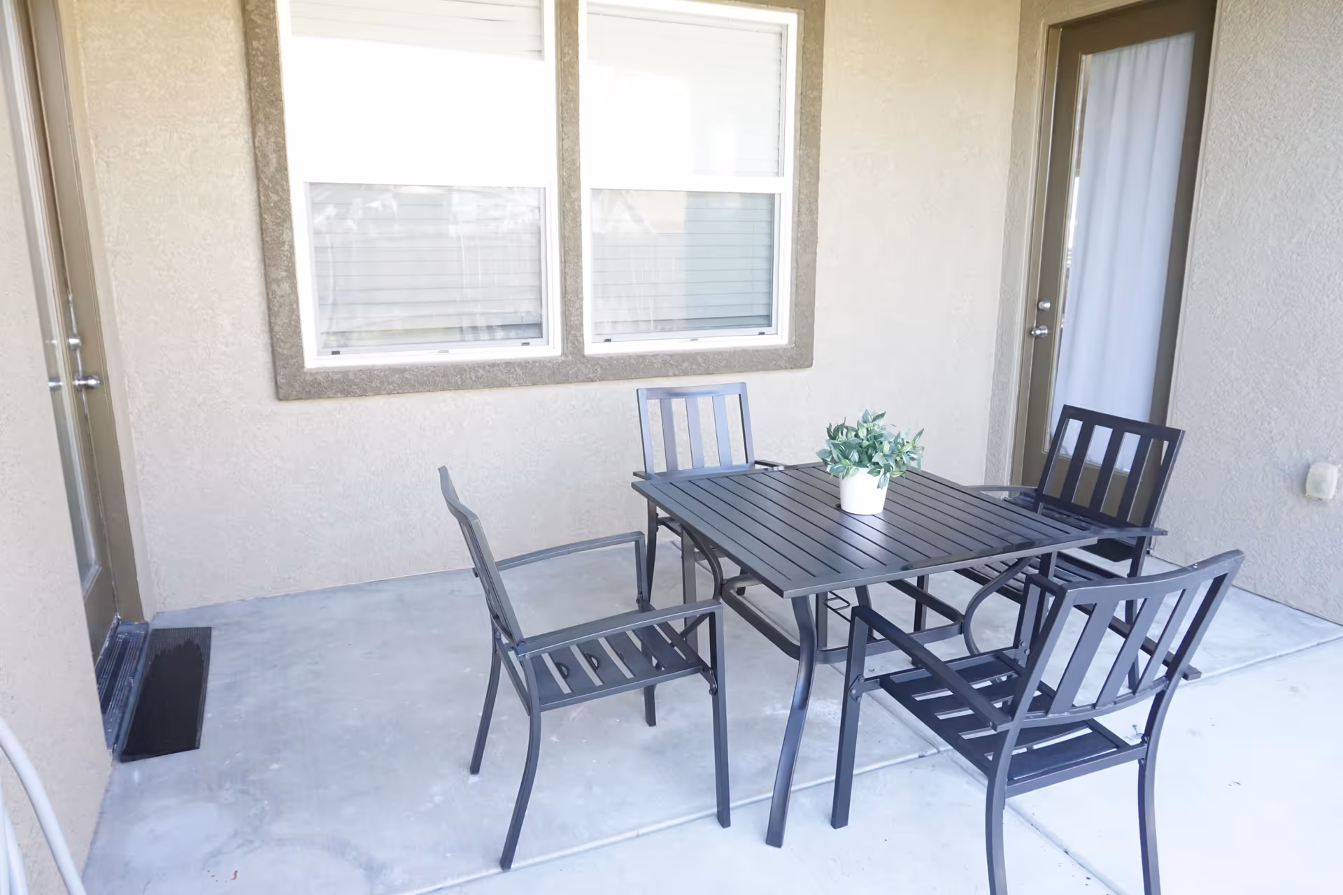Outdoor patio with a metal table, four chairs, and a potted plant near the entrance.