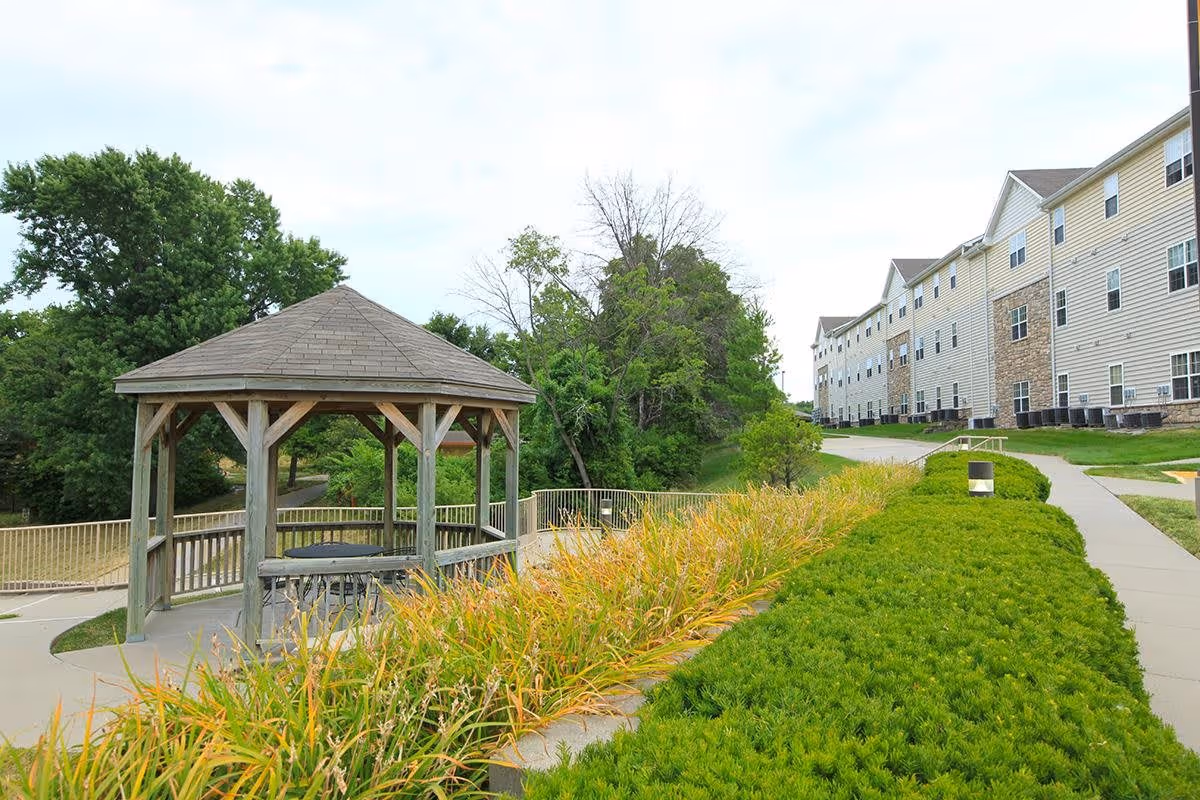 Outdoor view of a senior living facility with a wooden gazebo surrounded by greenery and a paved walkway. The building is visible on the right side with multiple windows and air conditioning units. Trees and bushes are present in the background.