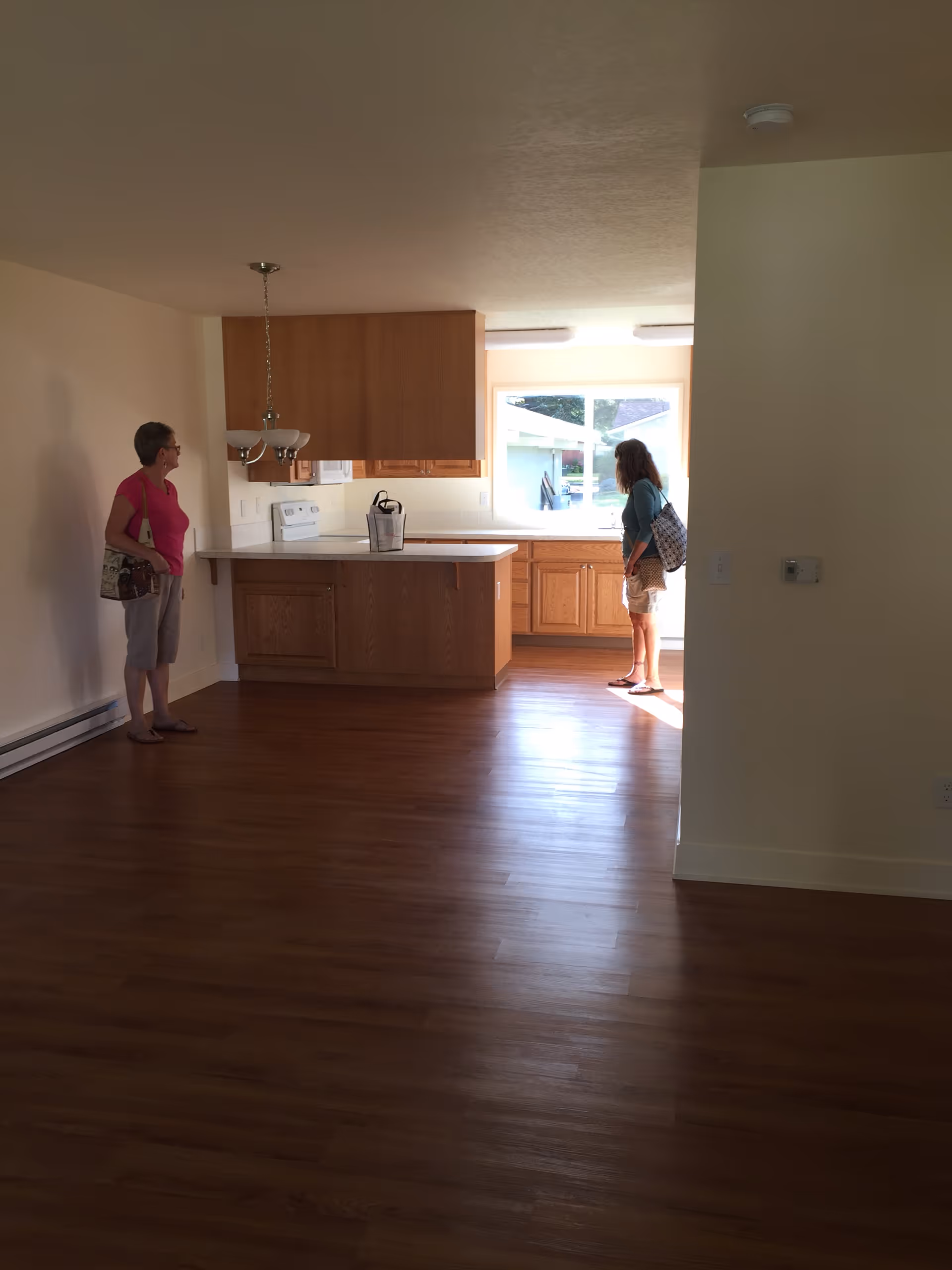 Interior view of a room with wooden flooring and a kitchen area in the background. Two women are standing in the room, one near the kitchen counter and the other near the wall. The kitchen has wooden cabinets, a stove, and a window letting in natural light.
