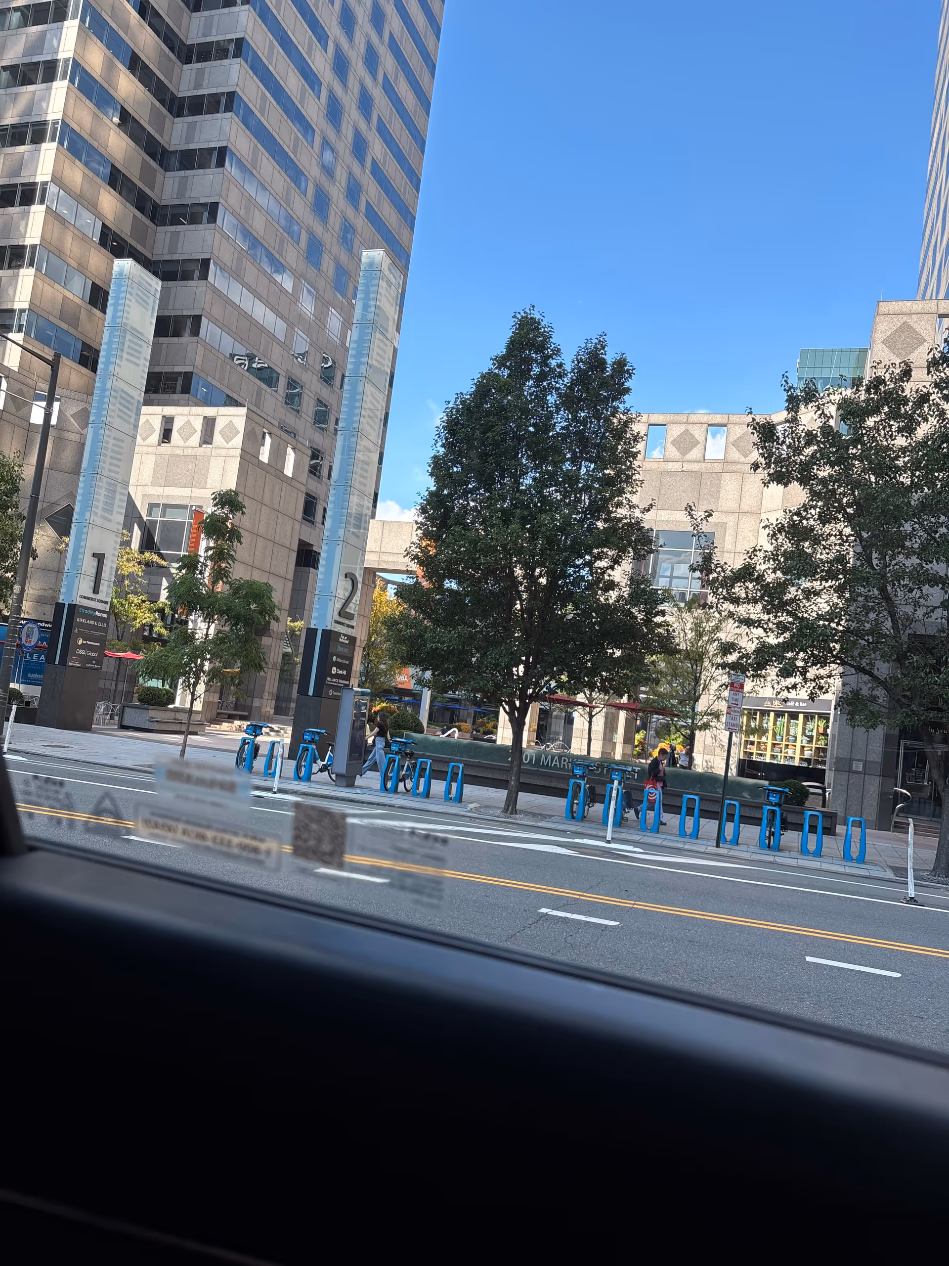 Urban street scene with tall modern buildings, trees, and blue bike racks along the sidewalk.