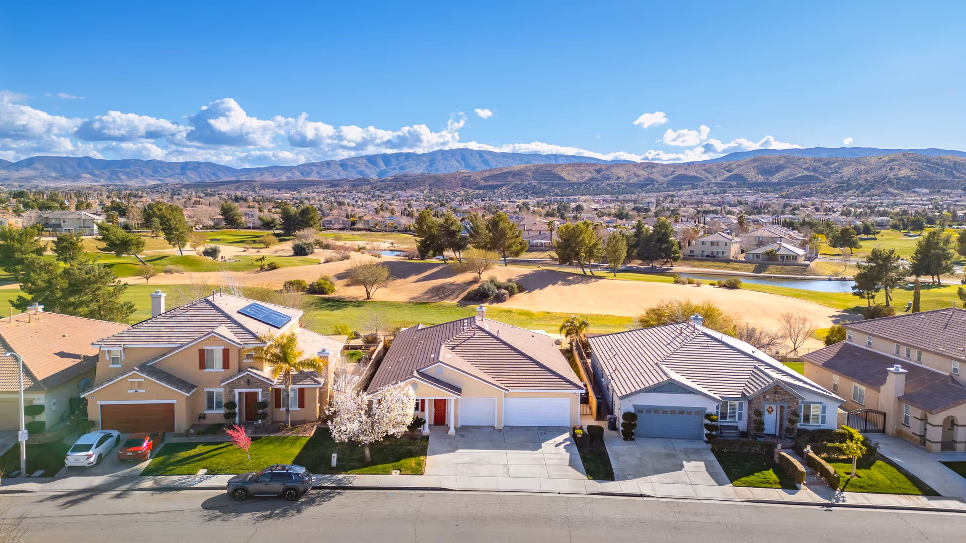 Row of suburban houses with manicured lawns along a street, hills and blue sky in background.