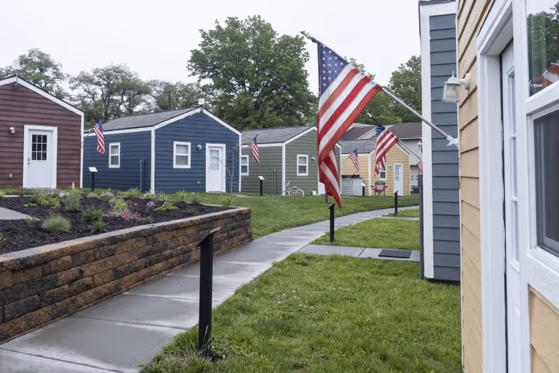 Colorful small cottages line a walkway outside Living Well Community Home with American flags.