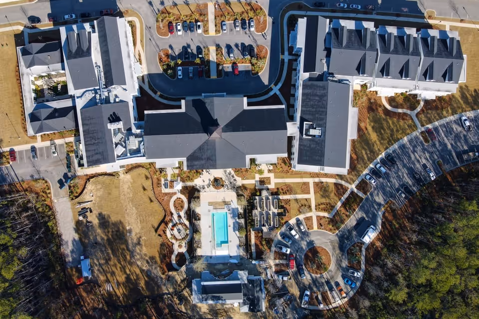Aerial view of Merrill Gardens at Carolina Park showing multiple buildings arranged around a central courtyard with a swimming pool, surrounded by parking lots and landscaped areas with trees and pathways.