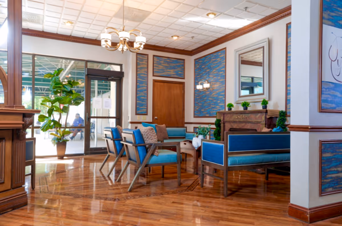 Bright furnished lobby with blue-upholstered chairs and sofas, wooden floors, a potted plant, and glass entry doors.