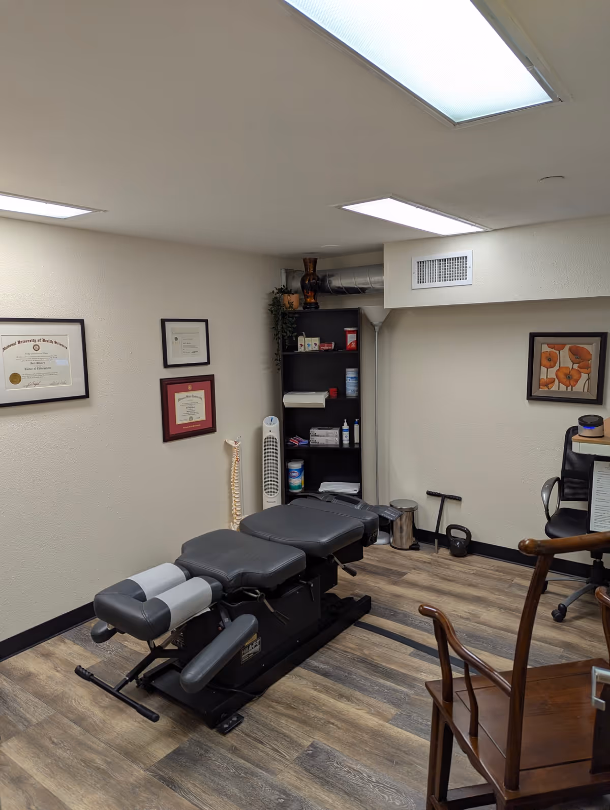 Chiropractic treatment table in a small therapy room with shelves and framed certificates on the wall.
