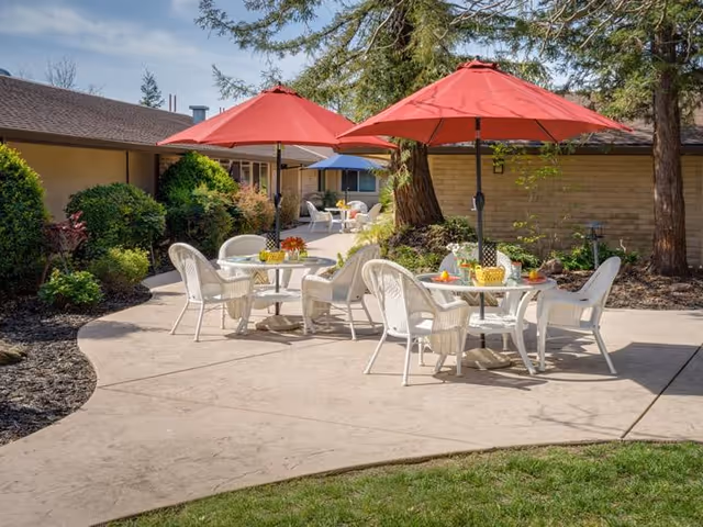 Sunlit courtyard with white patio tables and chairs under red umbrellas beside landscaped paths and a low brick building.