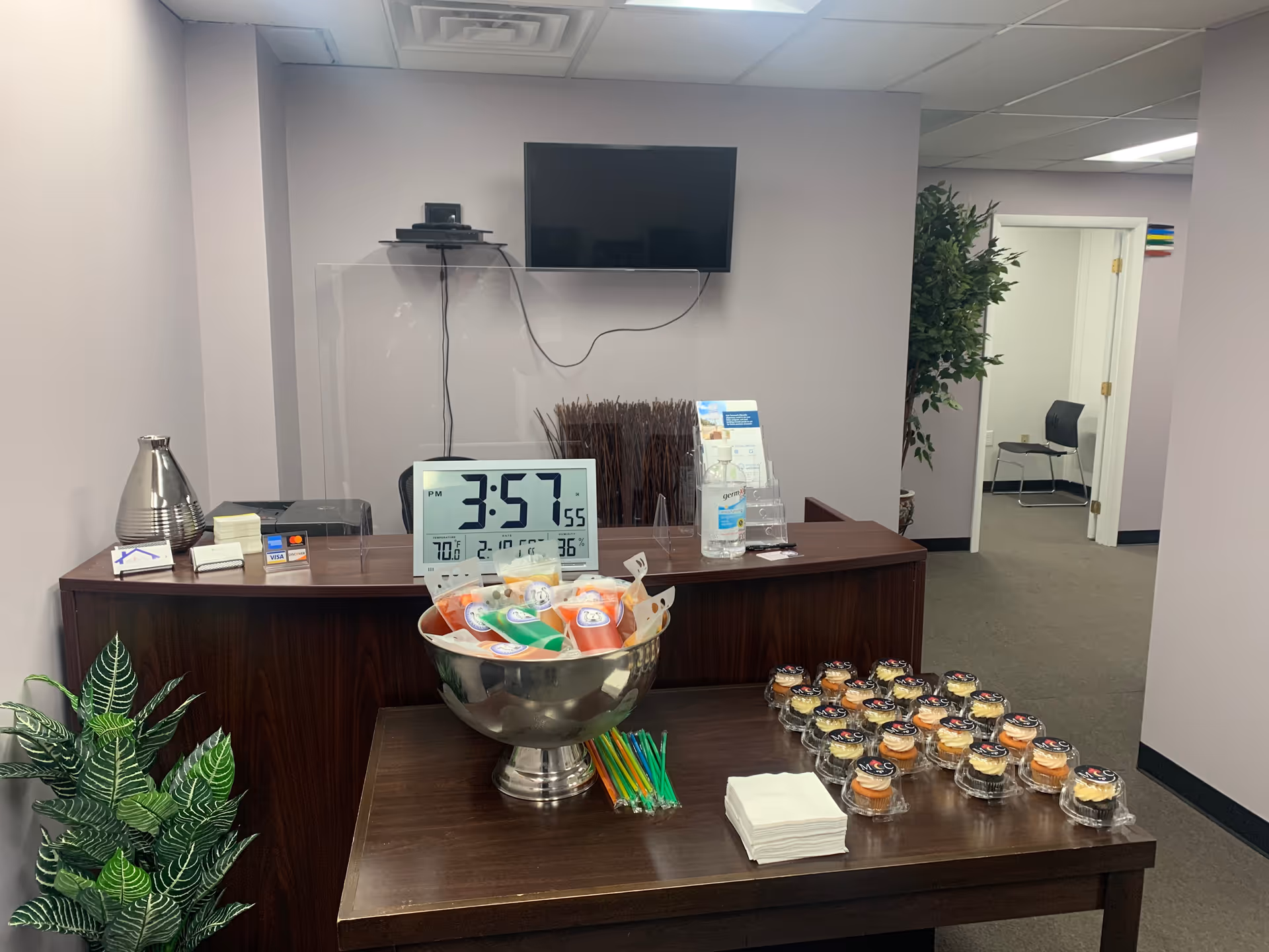Reception area with a wooden desk, wall mounted TV, cupcakes, clock, and plants.