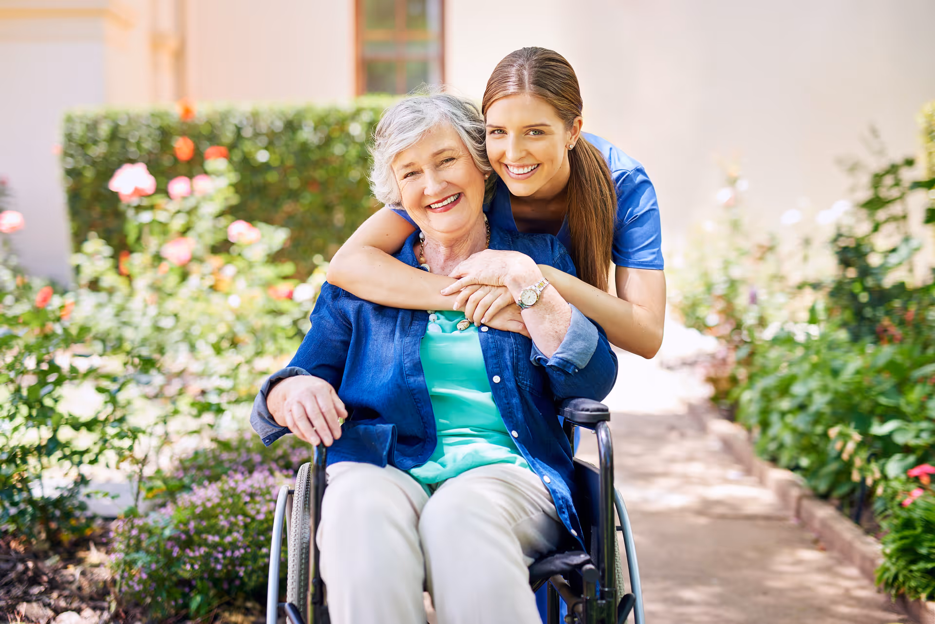 Young caregiver hugs a smiling elderly woman in a wheelchair outdoors in a garden.