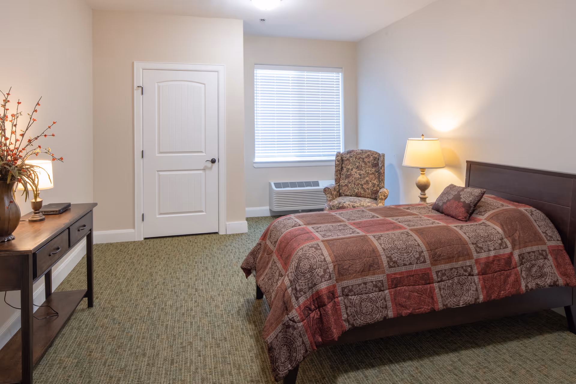 A furnished bedroom with bed, chair, dresser, lamp, window, door, and patterned quilt.