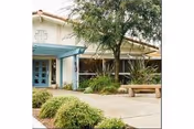 Exterior view of Pacific Coast Manor entrance with walkway, tree, and bench.