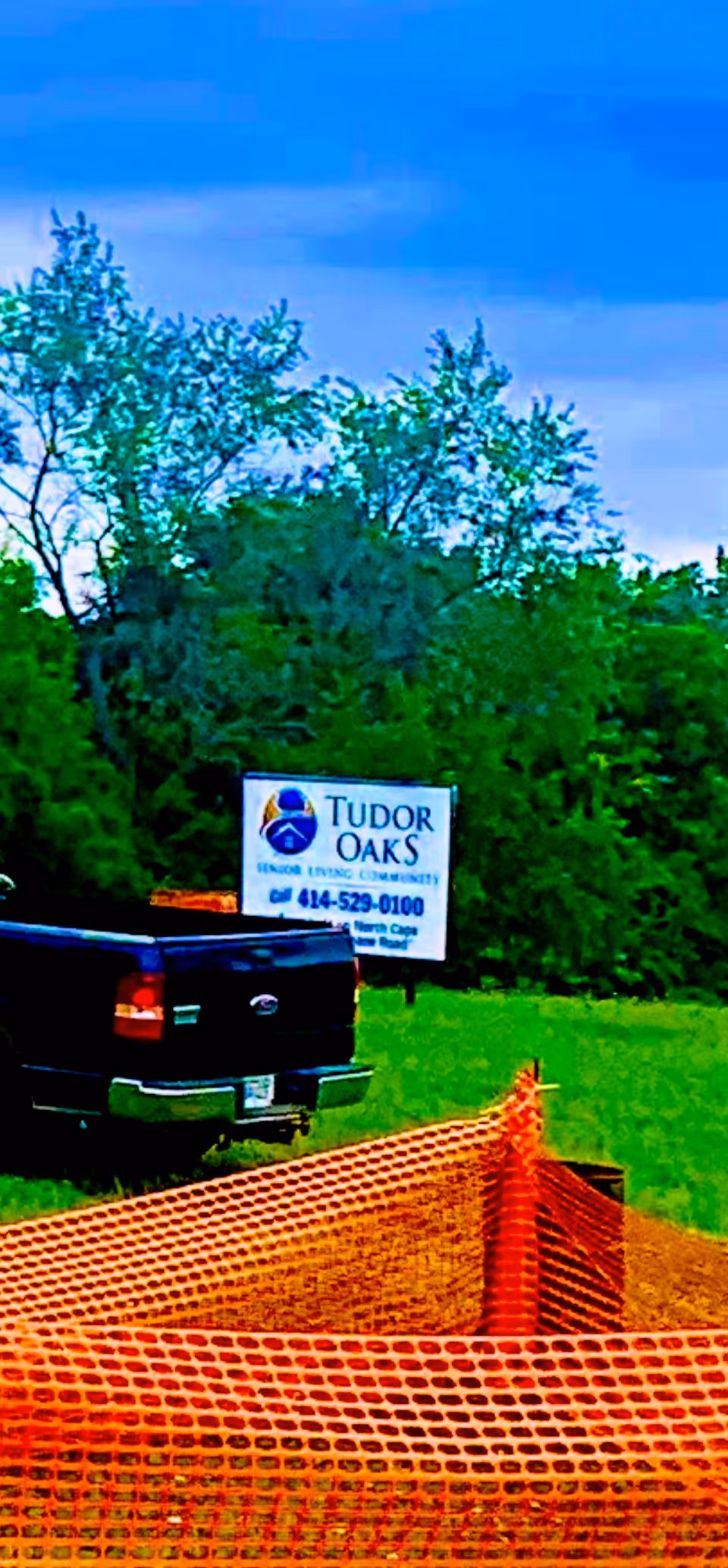 Outdoor scene featuring Tudor Oaks sign, grassy area, trees, a black pickup truck, and orange fencing.