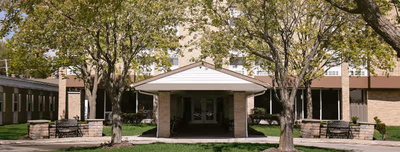Covered main entrance of Riverside Residence with a driveway, two trees, benches, and a brick building facade.