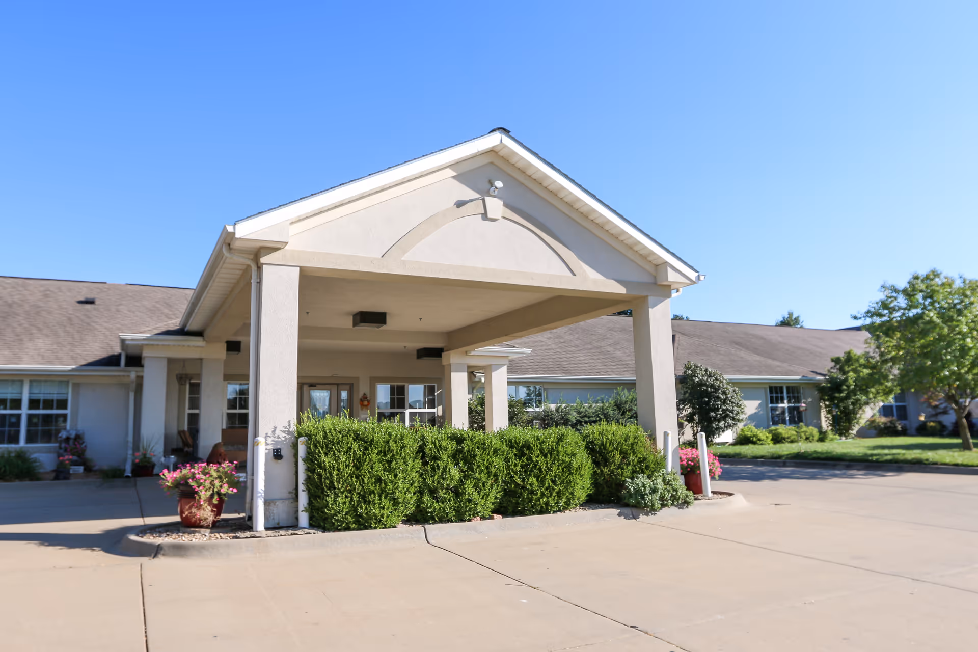 Exterior view of a senior living facility entrance with a covered portico and greenery.