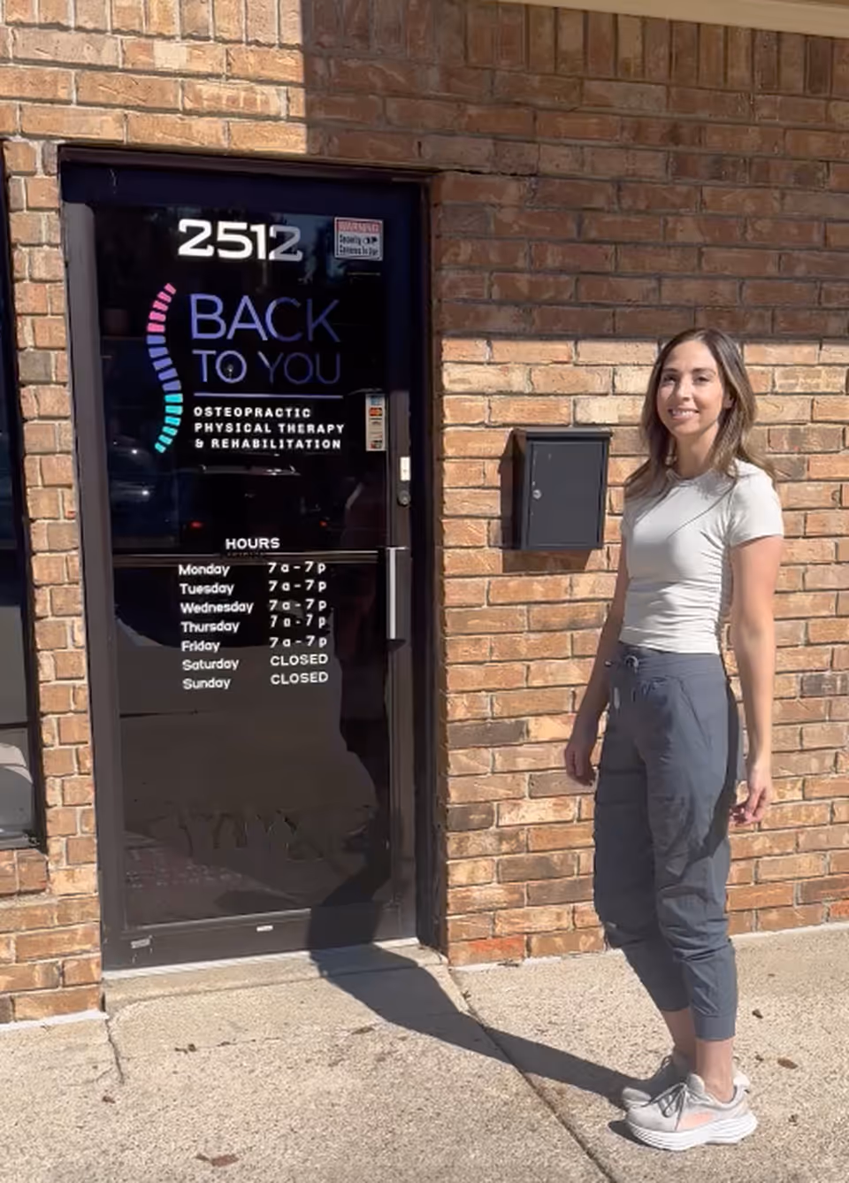 A woman stands outside a brick building next to a glass door with a therapy sign.