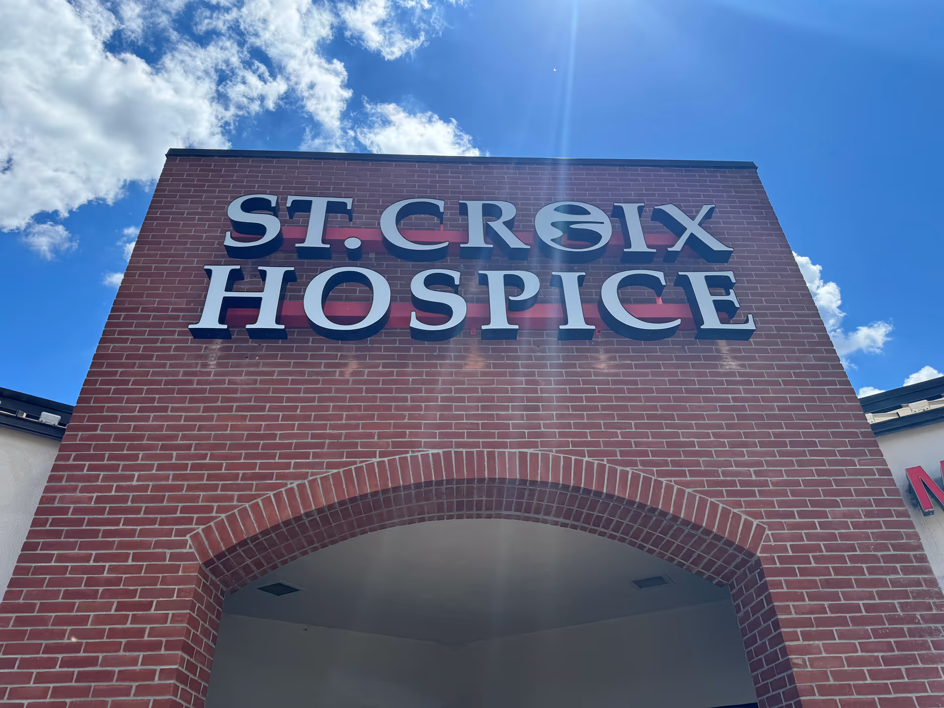 Brick building front with large St. Croix Hospice sign above an arched entrance, blue sky backdrop.