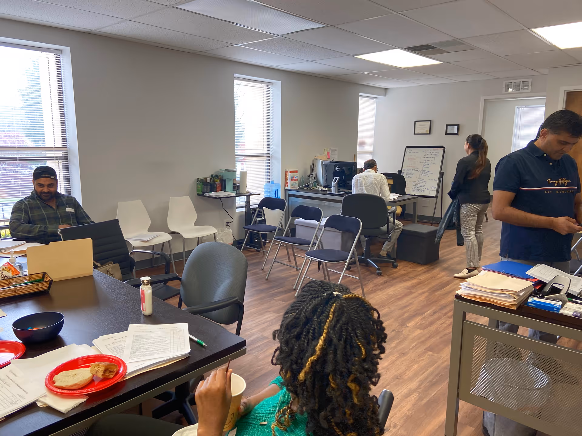 A communal interior room with desks, chairs, and staff assisting residents.