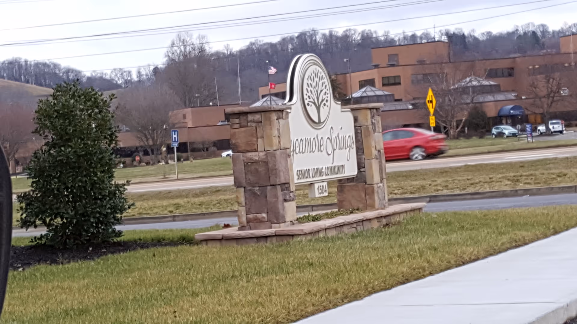 Entrance sign reads Sycamore Springs Senior Living Community with stone pillars and a building in background.