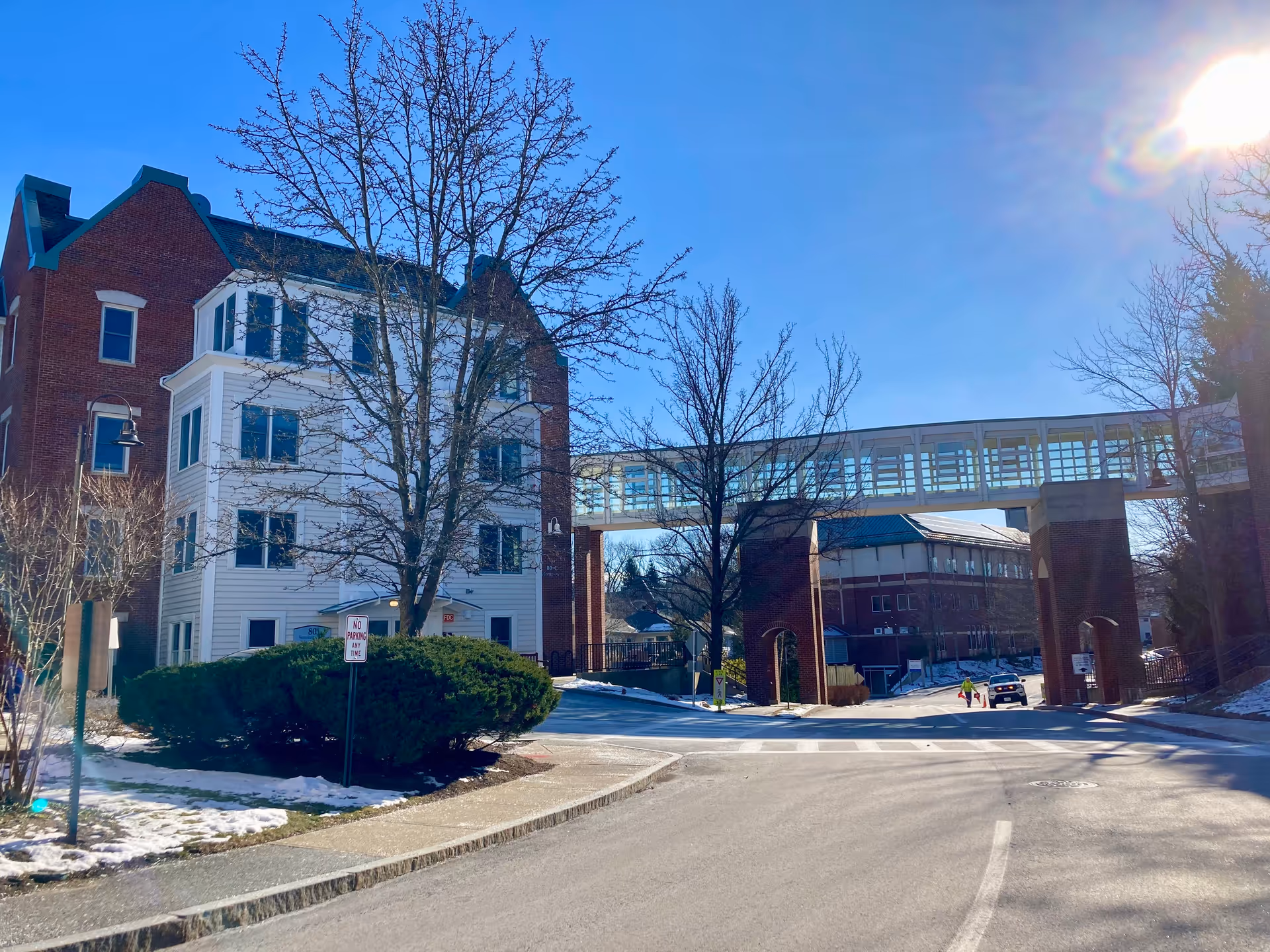 Exterior view of a senior living facility with a brick and white building on the left and a covered pedestrian bridge connecting two buildings. There are leafless trees, some snow on the ground, a road with a 'No Parking Any Time' sign, and a bright sun in the clear blue sky.